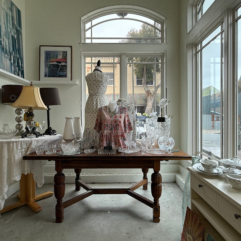 Interior of a store with a wooden table displaying glassware and a mannequin, large windows showing an outdoor view.